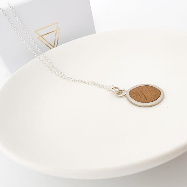 Collezione XXI Oak Pebble Necklace displayed on white background. Laid over a white dish. Long fine silver chain with silver and oak pendant. Vannucchi white packaging at back of image, with gold logo.