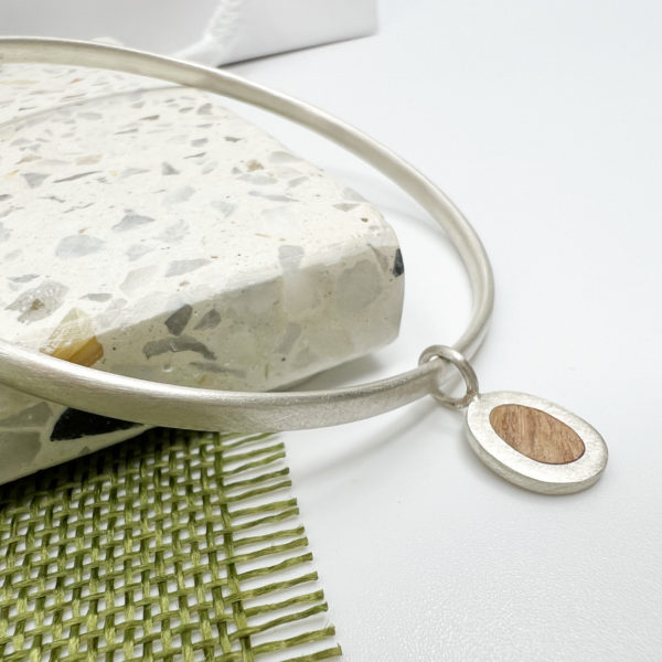 Silver collezione xxi pebble bangle displayed over a pale terrazzo tile on olive tree fabric and white background. Bangle is thin silver hallmarked, with a small oak wood and silver circular charm