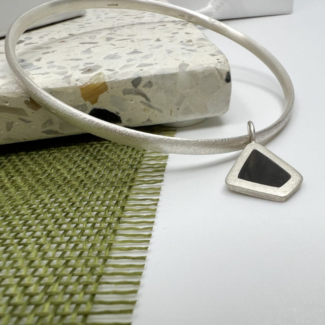 Silver collezione xxi black diamond bangle displayed over a pale terrazzo tile on olive tree fabric and white background. Bangle is thin silver hallmarked, with a small black wood wood and silver irregular diamond shaped charm
