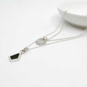 Collezione XXI Black Diamond Necklace. Two Silver pendants on fine silver chains laid over the edge of a white dish. One Pendant is small, round and textured. The larger pendant is in the shape of an irregular diamond and has a dark wood inlay.