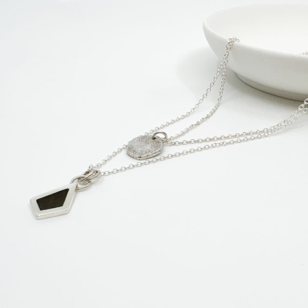 Collezione XXI Black Diamond Necklace. Two Silver pendants on fine silver chains laid over the edge of a white dish. One Pendant is small, round and textured. The larger pendant is in the shape of an irregular diamond and has a dark wood inlay.