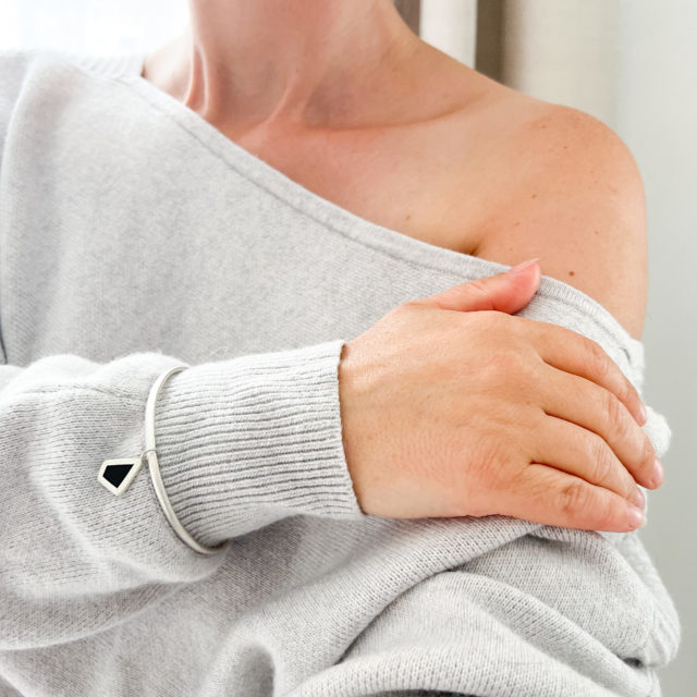 Model wears the Collezione XXI Black Diamond Bangle over sleeve of grey jumper. Holding arm across the body they showcase the thin silver textured bangle with small black wood and silver irregular diamond shaped charm