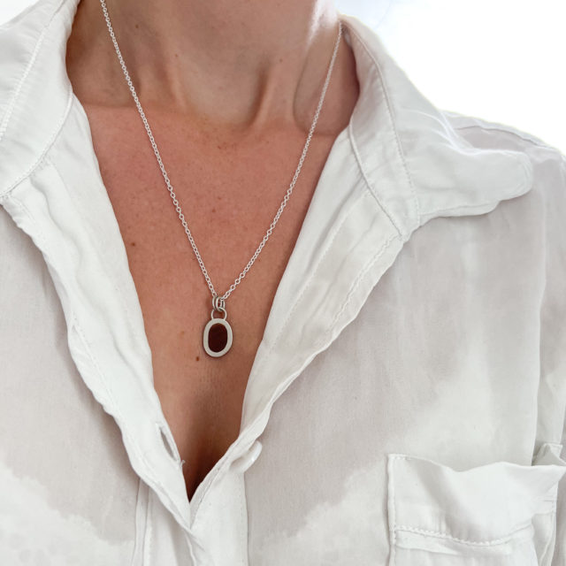 Model wears Collezione XXI Wood Pebble Necklace. The model wears a white silk shirt slightly open to expose the necklace on long fine silver chain. Light red coloured wood inlay and silver pebble shaped small pendant.