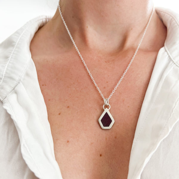 Model wears Collezione XXI Purple Heart Necklace. White shirt is slightly open with fine silver chain necklace, Pendant pin necklace is an irregular pentagon with purple wood inlay.