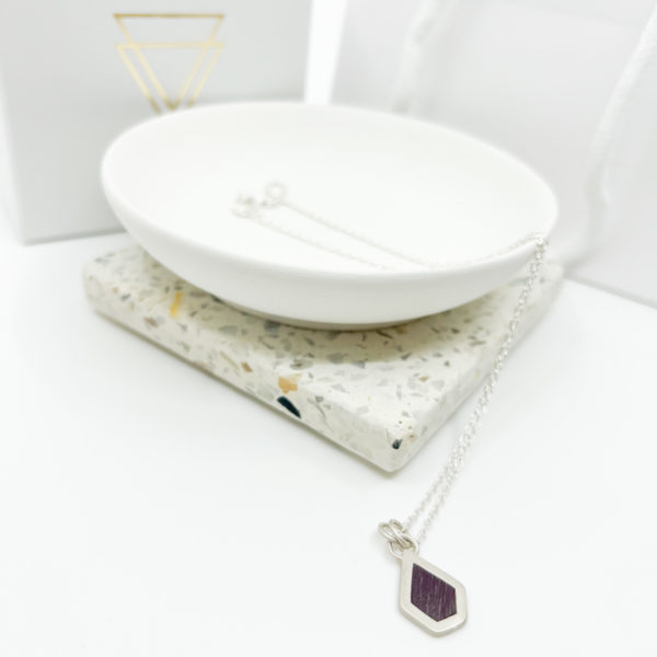 Collezione XXI Purple Heart necklace. Long fine silver chain laid over the edge of a white dish. Small silver, irregular pentagon pendant has a purple heart wood inlay. Vannucchi packaging in the background with gold logo.