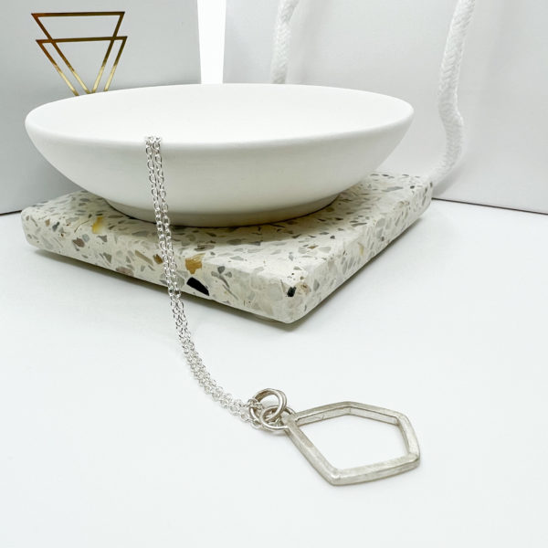 Collezione XXI Large Stone Necklace. Irregular geometric shaped pendant made from silver square wire on fine silver chain. Laid over the edge of a white dish. White Vannucchi packaging in the background with gold logo.