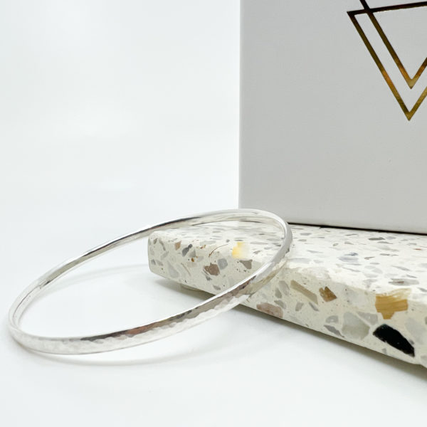 The Collezione XXI Hammered Bangle is a thin silver bangle, laid over a pale terrazzo tile in a white background with white Vannucchi packaging behind.