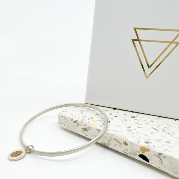 The Collezione XXI Pebble Bangle is a thin silver bangle with a small oak wood and silver circular charm. Laid over a pale terrazzo tile in a white background with white Vannucchi packaging behind.