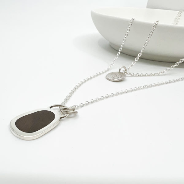 Collezione xxi multi layered necklace. Two Silver pendants on fine silver chains laid over the edge of a white dish. One Pendant is small, round and textured. The larger pendant has a dark wood inlay.