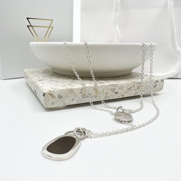 Collezione XXI Multi Layered Necklace. Two Silver pendants on fine silver chains laid over the edge of a white dish. One Pendant is small, round and textured. The larger pendant has a dark wood inlay. Vannucchi packaging in the background with gold logo.