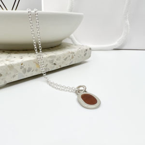 Collezione XXI Wood Pebble Necklace. Long fine silver chain laid over the edge of a white dish. Small pebble shaped pendant has a light red wood inlay. Vannucchi packaging in the background with gold logo.