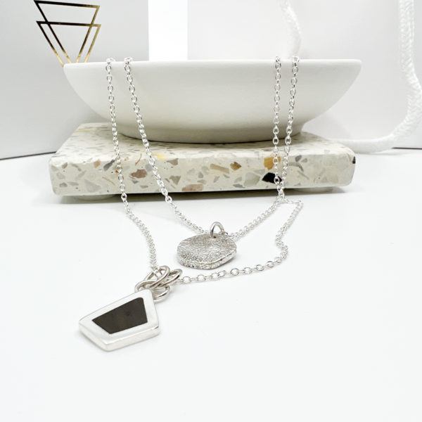 Collezione XXI Black Diamond Necklace. Two Silver pendants on fine silver chains laid over the edge of a white dish. One Pendant is small, round and textured. The larger irregular diamond pendant has a black wood inlay. Vannucchi packaging in the background with gold logo.