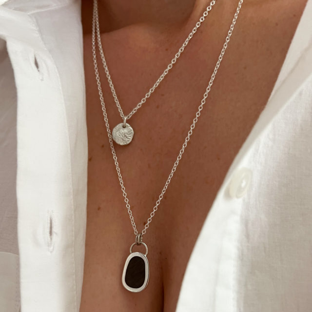 Model wears a white shirt with Collezione xxi multi layered fine chain necklace. Two strands. Shorter chain has small, round textured pendant. Longer chain has a dark wood and silver pendant attached.